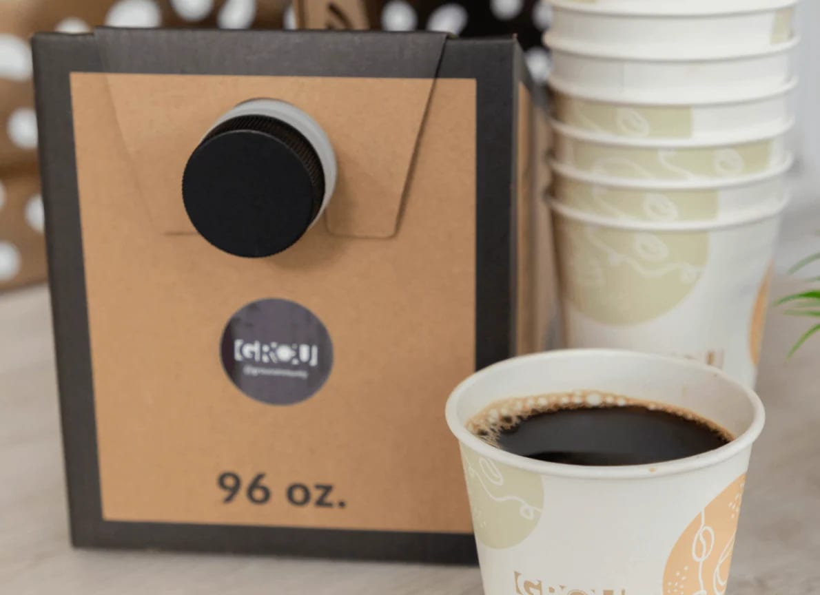 GROU 96 oz Coffee Box with a spigot next to a stack of GROU paper cups and a poured cup of black coffee