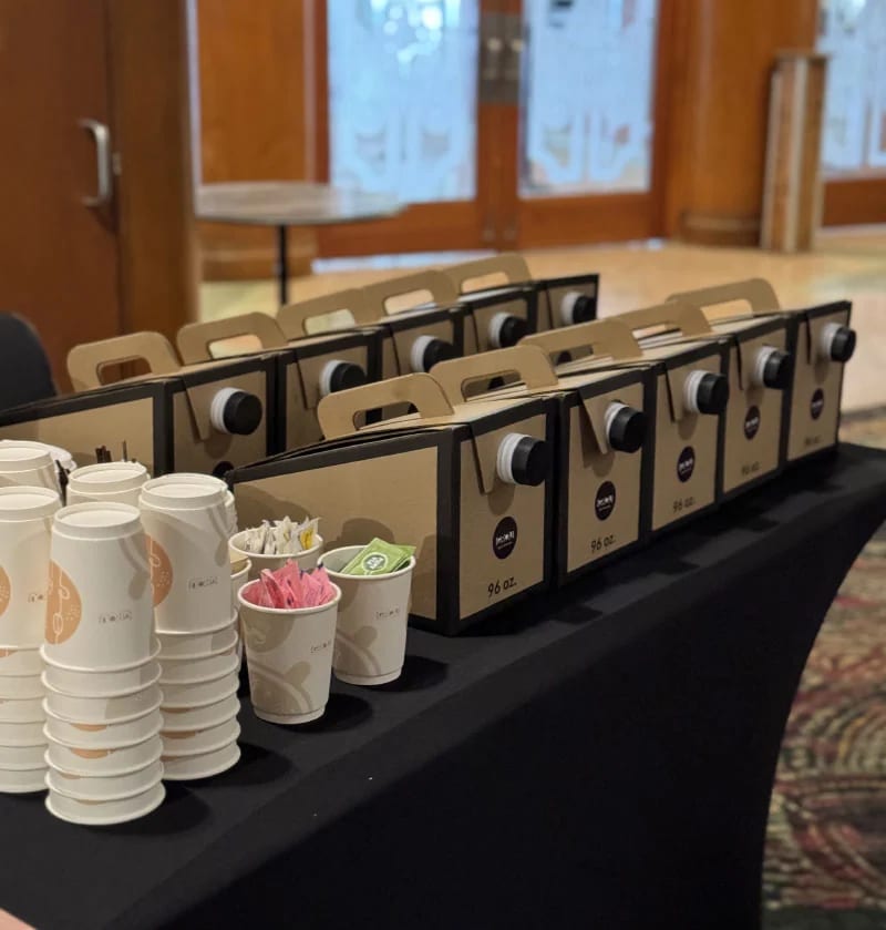 Ten GROU 96 oz coffee boxes lined up on a catering table with stacks of cups, sweeteners and tea bags